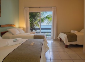 Habitación de hotel con dos camas, un balcón con vista a palmeras y al mar, y un cabecero de madera.