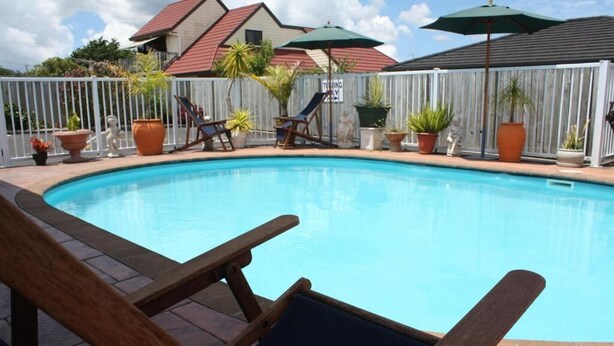 Piscina com espreguiçadeiras e guarda-sóis, rodeada por um cercado branco e plantas em vasos.