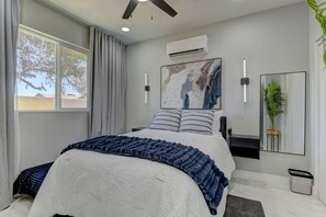 Une chambre moderne avec un lit, un ventilateur de plafond, un appareil de climatisation, une grande fenêtre avec des rideaux, un climatiseur mural, un tableau encadré, un miroir et une plante en pot.