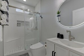 Une salle de bain moderne avec une douche à parois de verre, un lavabo blanc doté d’un distributeur de savon noir et un miroir rond.