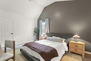 Un dormitorio con techo inclinado, una cama con ropa de cama blanca, una mesita de noche con una lámpara y una silla.