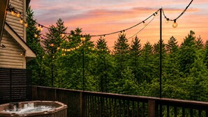Eine Holzterrasse mit Whirlpool, umgeben von einem Holzzaun, mit Lichterketten vor einer Baumkulisse und einem Sonnenuntergangshimmel.