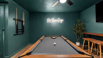 A pool table in a room with teal walls, a neon sign, a ceiling fan, and a potted plant.