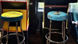Zwei Spielautomaten, einer mit der Aufschrift 'PAC-MAN' und der andere mit 'THE SIMPSONS', jeweils mit Sitzhocker davor.