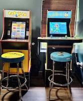 Dos máquinas de arcade, una con la etiqueta 'PAC-MAN' y la otra con 'THE SIMPSONS', con taburetes frente a cada una.