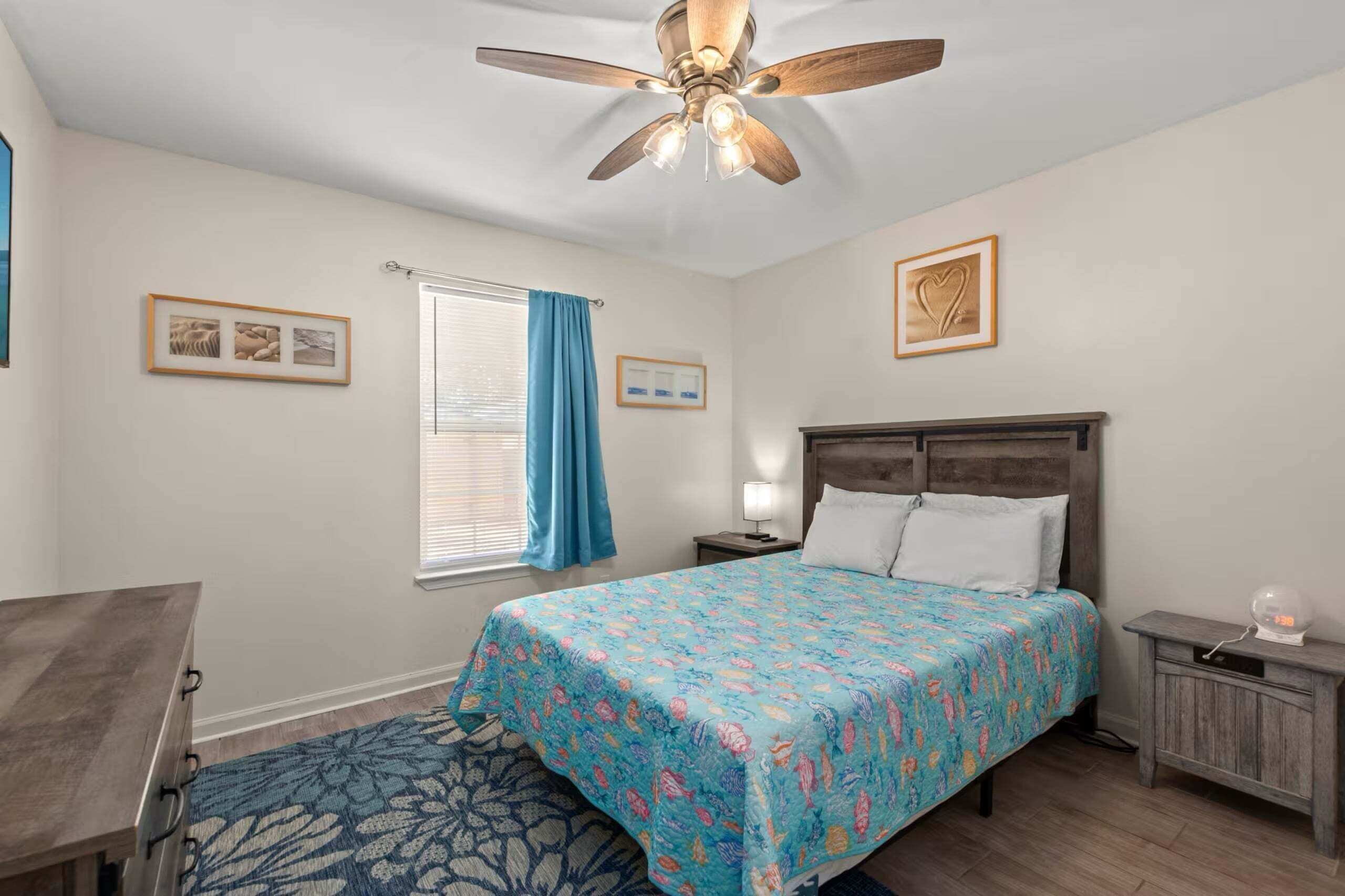 Quarto com cama, mesa de cabeceira, escrivaninha, cadeira, ventilador de teto e janela com cortinas.