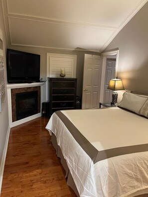 A bedroom with a large bed, a television above a fireplace, a door, and a nightstand with a lamp.