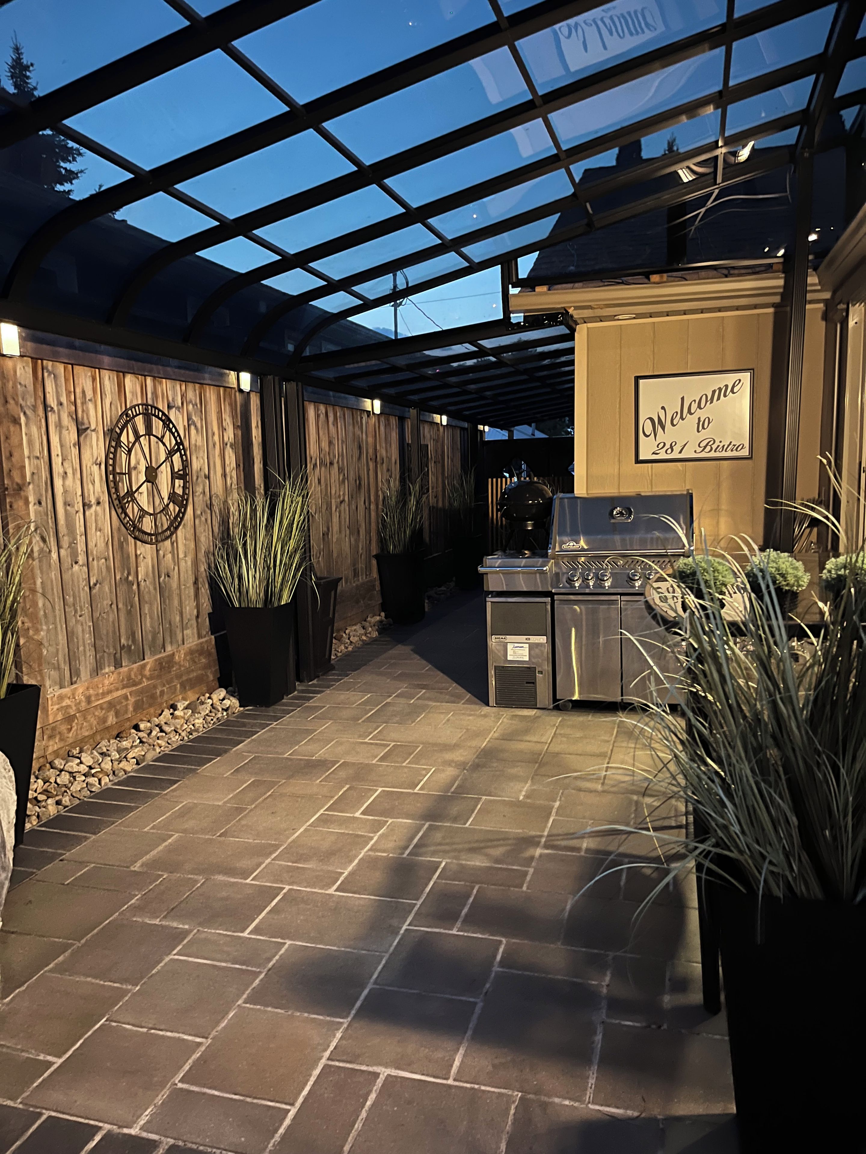 Un patio exterior techado con una parrilla, plantas en macetas y un cartel de bienvenida.
