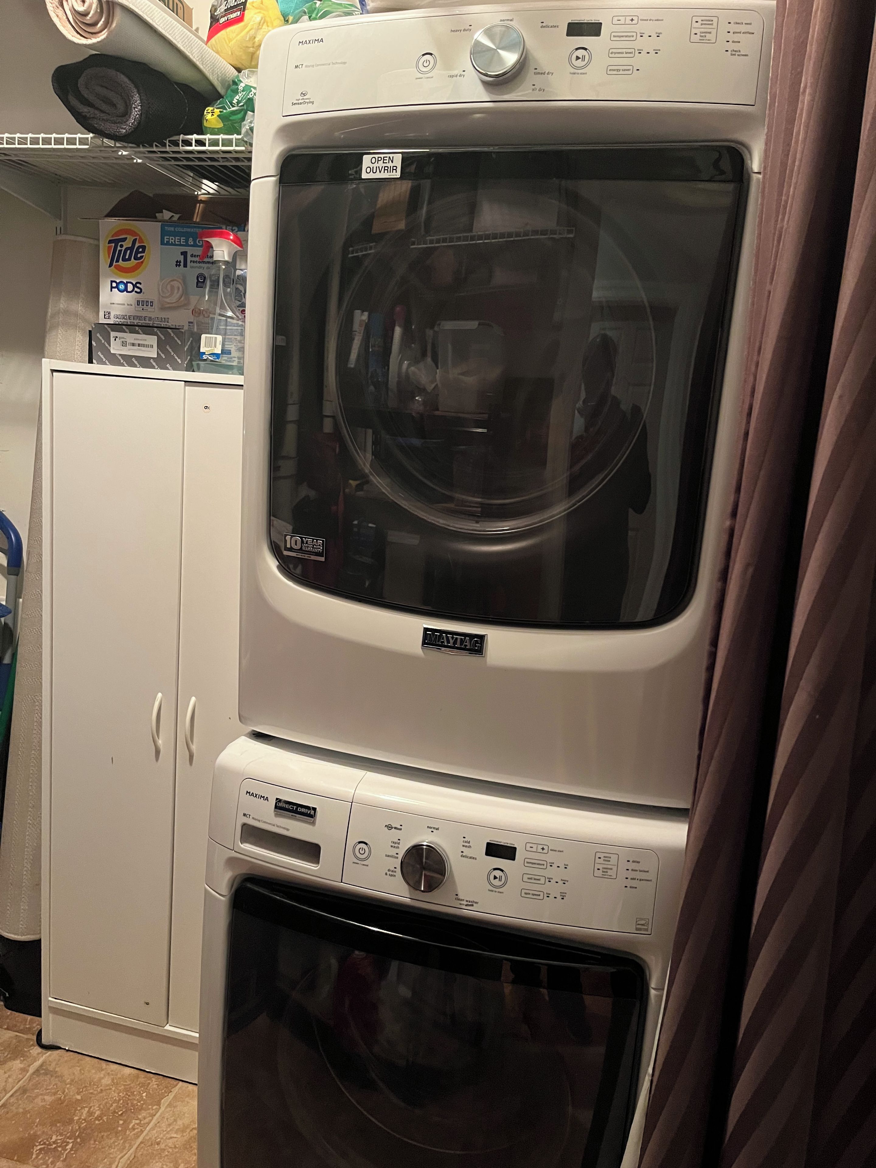 Lavadora y secadora Maytag de carga frontal con panel de control, ubicadas en un cuarto de lavado con un gabinete blanco y un estante arriba.
