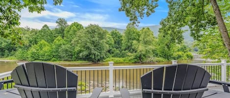 Una terraza con dos sillas Adirondack orientadas hacia un río, rodeada de árboles y una baranda blanca.