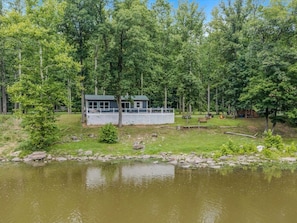 A lakeside cabin with a deck and outdoor seating area, surrounded by trees and a calm body of water. - Welcome to the river - River, Wifi, Deck, Fire Ring (Great Cacapon)