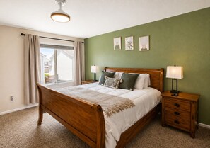 Une chambre avec un lit en bois, une table de chevet avec une lampe, une fenêtre avec des rideaux et un mur vert orné de trois tableaux encadrés.