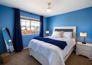 A bedroom with a bed, two bedside tables, a window with curtains, a ceiling light, and a view of houses outside. - 3BR Home w/ Private Cinema • BBQ • Home Office • 10 minutes from Airport (Denver)