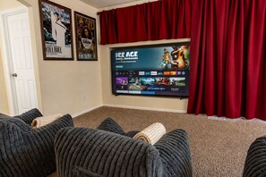 Une salle de cinéma privée équipée d’un grand téléviseur à écran plat diffusant un film, d’un rideau rouge et d’un sol recouvert de moquette.