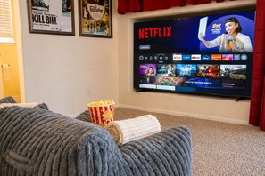 A living room with a large flat-screen TV displaying a Netflix interface, a popcorn bucket, and a cozy grey sofa. - 3BR Home w/ Private Cinema • BBQ • Home Office • 10 minutes from Airport (Denver)