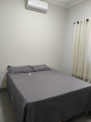 A single bed with a grey bedspread and two pillows, an air conditioning unit on the wall, and a window with white curtains.