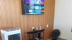 A room with a bed, a standing heater, a TV mounted on a wooden wall, and a fan on the floor.