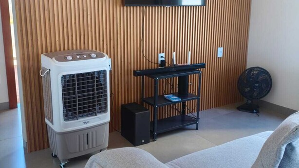 A room with a flat-screen TV mounted on a wooden wall, a white portable air cooler, a black speaker, and a black fan on the floor.