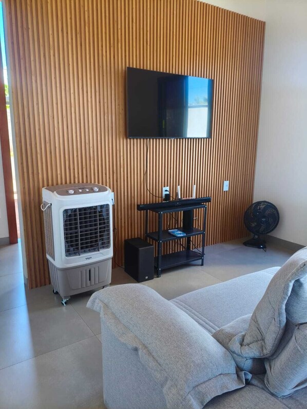 Une pièce avec un téléviseur à écran plat fixé à un mur en bois, un climatiseur portatif blanc, un haut-parleur noir et un ventilateur noir posé sur le sol.