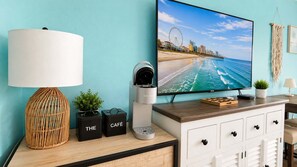 A room with a coffee machine, a television displaying a beach scene, a lamp, and storage boxes labeled 'THE' and 'CAFÉ'. - Endless Ocean Views | 15th Floor King Studio (Myrtle Beach)