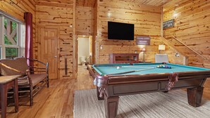 A wooden cabin interior with a pool table, a television mounted on the wall, and a sign welcoming guests to the cabin.