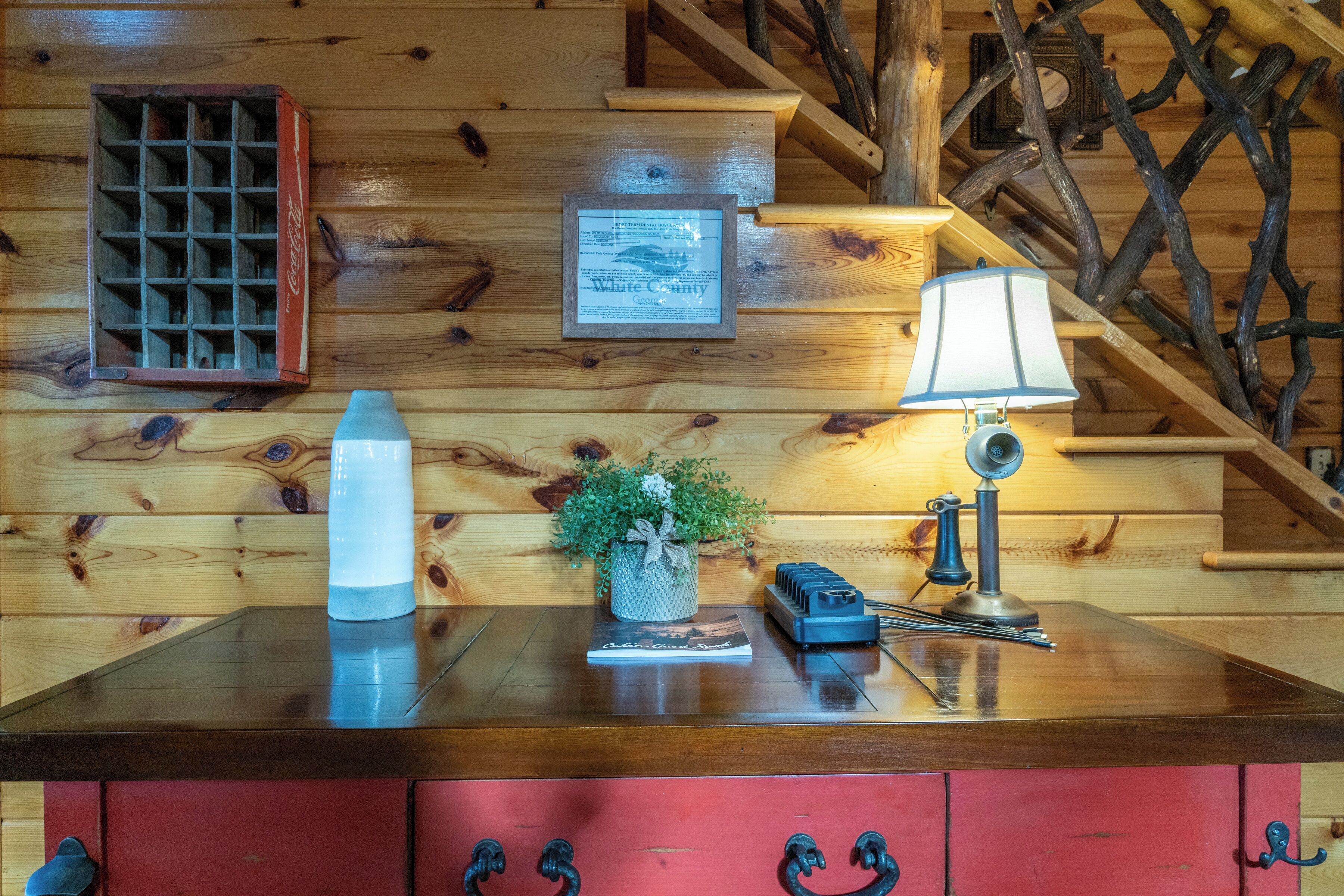 L’intérieur d’un chalet en bois, avec un escalier en bois, un réfrigérateur Coca-Cola, un certificat encadré, une lampe, une petite plante en pot et un buffet rouge avec des poignées en métal.