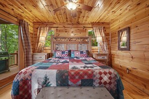 Ein Schlafzimmer in einer Blockhütte mit einem großen Bett, zwei Nachttischen, einem Deckenventilator und einem Fenster mit Vorhängen.
