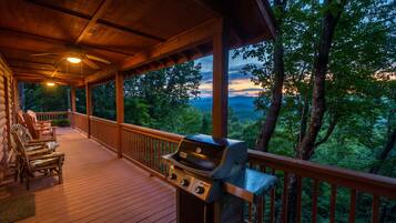 A covered outdoor deck with a grill, seating area, and a view of the surrounding forest.