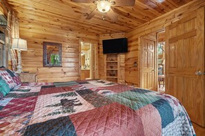 A wooden cabin bedroom with a large bed, a ceiling fan, a television, and wooden doors.