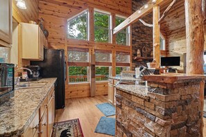 A cabin-style kitchen with wooden walls, large windows, a granite countertop, and a stone accent wall.