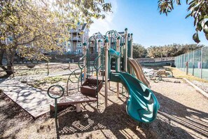A playground with a slide and climbing structures, surrounded by trees and a fence. - Barrier Island Station, Duck, NC - 2 Bedroom/Sleeps 8 (Duck)