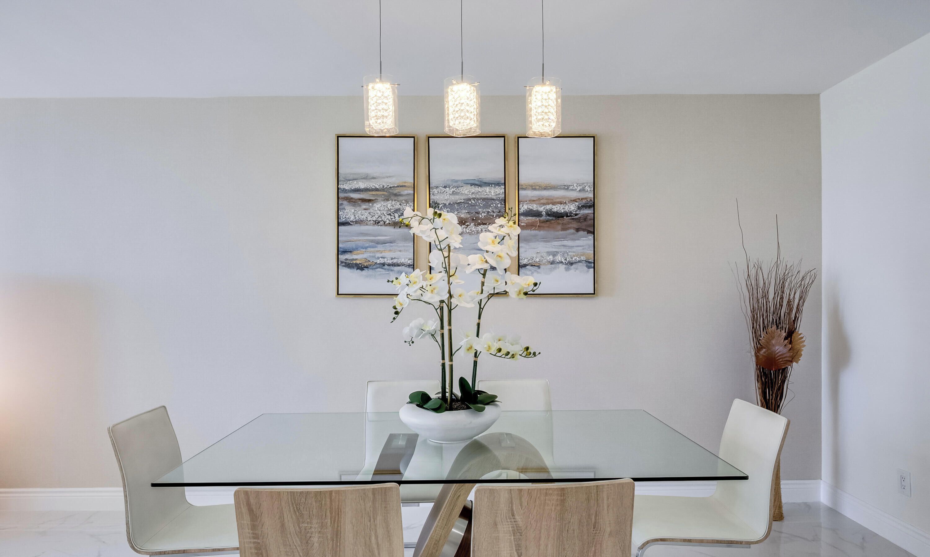 Un comedor con mesa de vidrio, sillas blancas, una planta de orquídeas en el centro y un cuadro de paisaje en la pared.