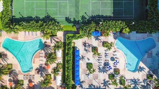Ett flygbildsbild av ett semesterområde med två stora pooler, en tennisbana och ett stort antal solstolar.