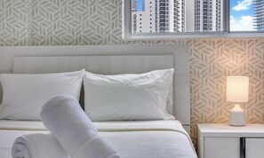 A neatly made bed with white linens, a bedside lamp, and a window with a view of tall buildings. - Oceanview 1BR Condo | Pool/Tennis/Beach (Sunny Isles Beach)