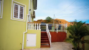A two-story building with a staircase leading to a balcony, a gated entrance, and a view of a hill in the background. - Experience the Caribbean sophistication at this Ultra-modern, 6 Bedroom En-suite (Plymouth)