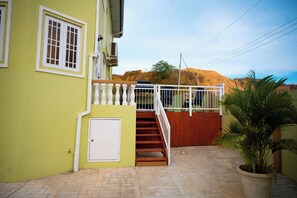 Ein zweistöckiges Gebäude mit einer Treppe, die zu einem Balkon führt, einem befestigten Eingang und Blick auf eine Hügelkulisse im Hintergrund.