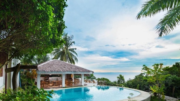 A pool area with a thatched-roof structure, surrounded by lush greenery and a view of the sea. - Fantasy Lodge (Samboan)