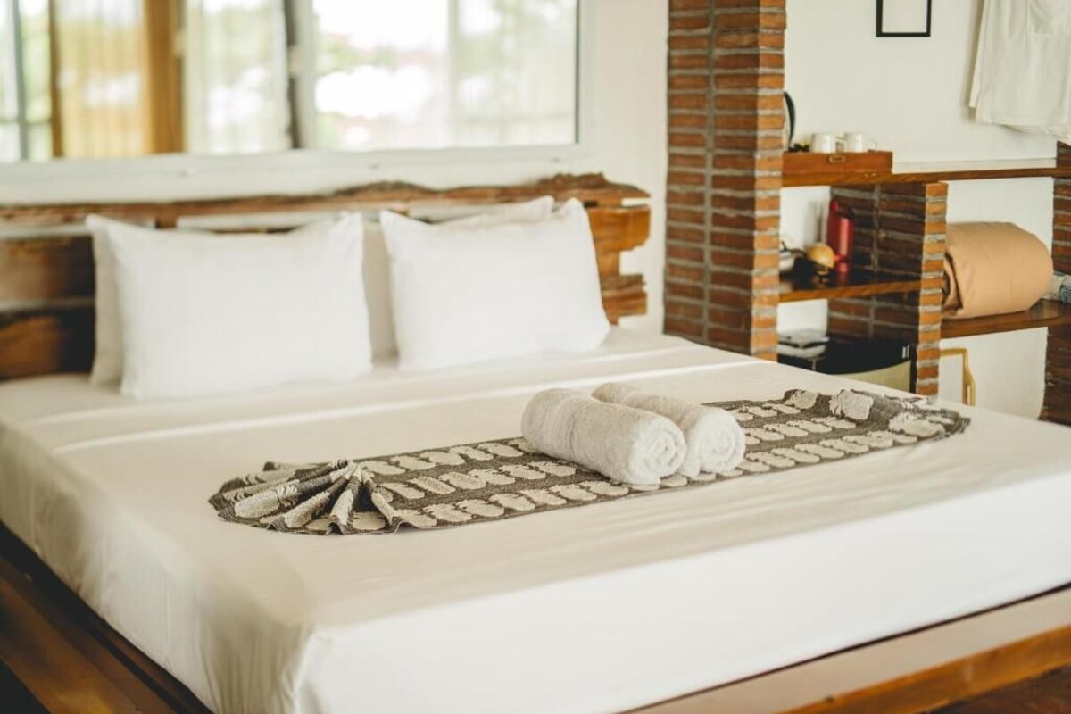 Una cama bien tendida con sábanas blancas y un plaid estampado, un cabecero de madera y una pared de ladrillo decorativa.