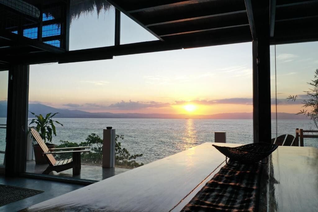 A room with a view of the sunset over the sea, a table, chairs, and a plant.