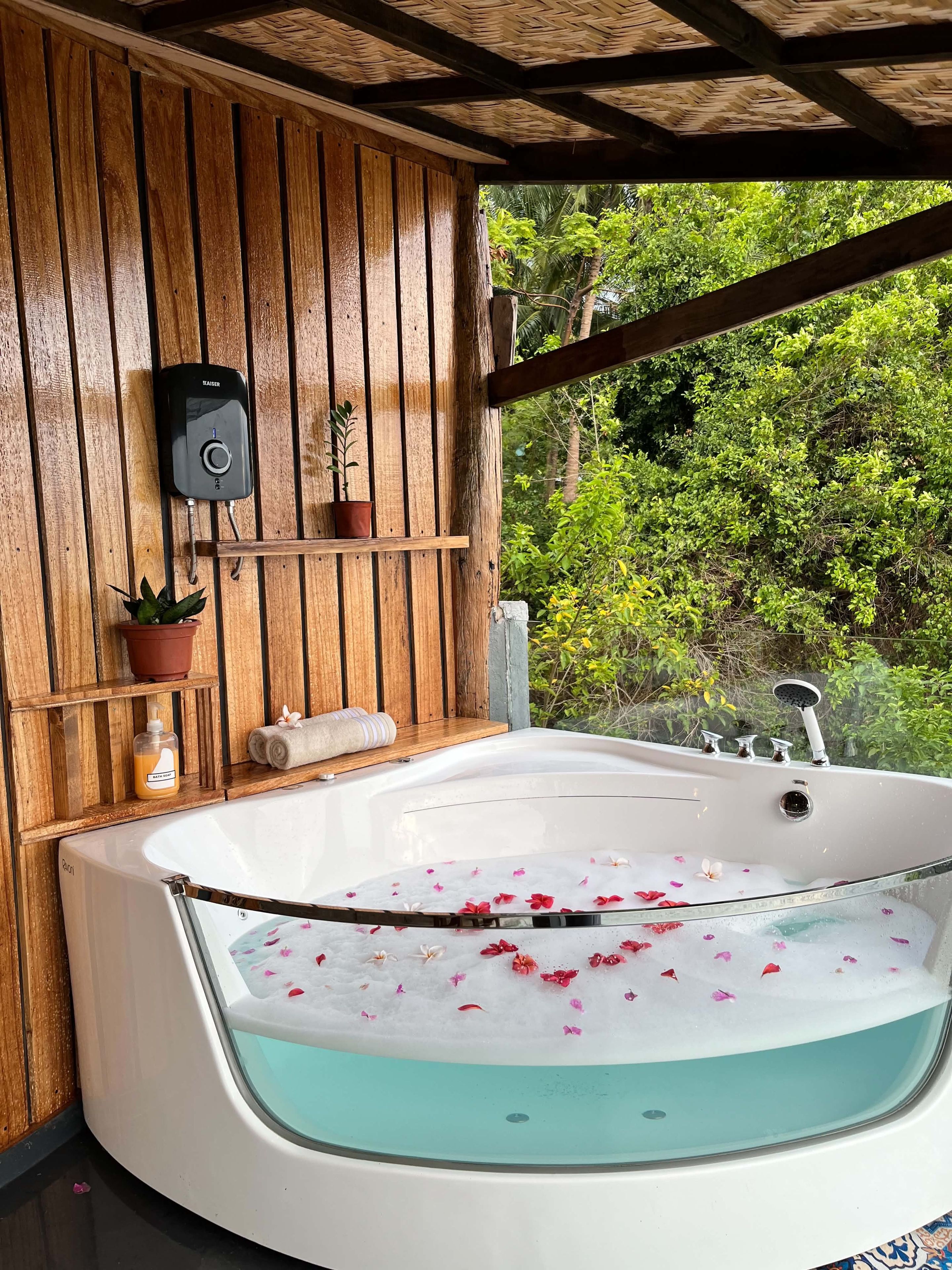 Zona de spa con bañera llena de agua y pétalos de rosa, pared de madera, calentador de agua instalado y una planta en maceta.