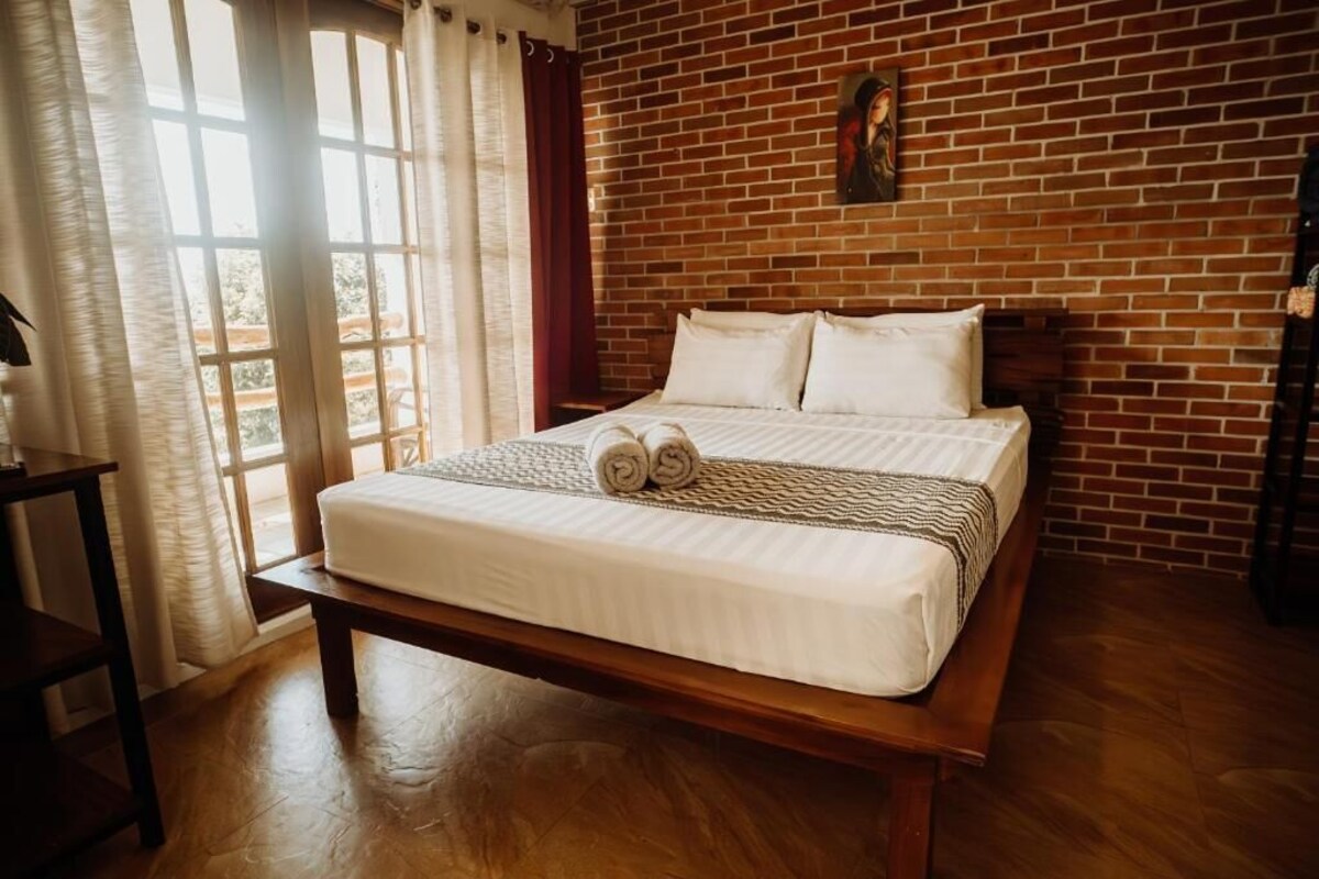 Una habitación con pared de ladrillo, cama con ropa de cama blanca, un banco y una ventana con cortinas.