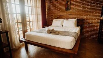 Een kamer met een bakstenen muur, een bed met witte beddengoed, een bankje en een raam met gordijnen.