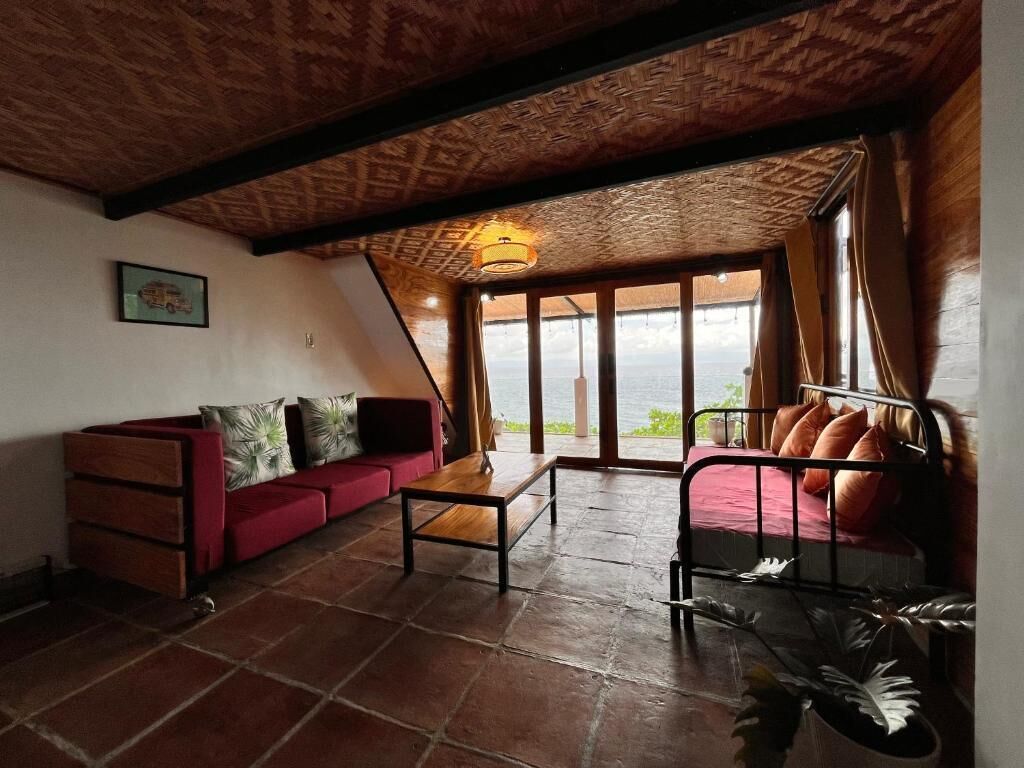 A room with a red sofa, a small table, and a metal bed frame. There is a large window with a view of the outdoors.