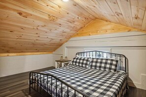 Ein Schlafzimmer mit schräg verlaufender Decke, einem Metallbettgestell, kariertem Bettzeug, einem kleinen Nachttisch mit zwei Lampen und einem Fenster.