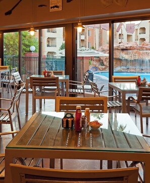 Un espace de restauration en plein air avec des tables et des chaises, une table en verre sur laquelle sont disposés des condiments, et une vue sur une piscine et des bâtiments.