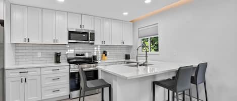 Cozinha moderna com armários brancos, ilha central com banquetas, eletrodomésticos de aço inoxidável e iluminação embutida no teto.