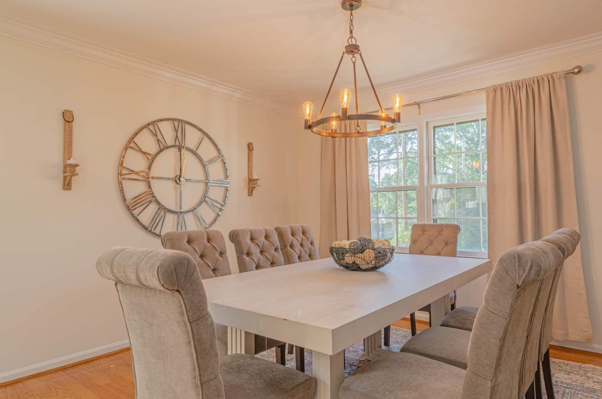 A dining room with a large table, six chairs, a wall clock, and a chandelier.