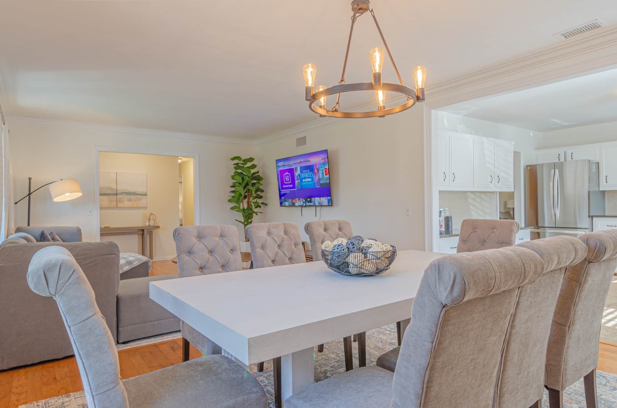 A modern dining area with a white table, tufted chairs, a chandelier, and a television on the wall.