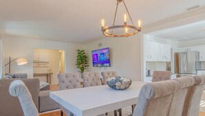 Uma sala de jantar moderna com mesa branca, cadeiras estofadas, um lustre e uma televisão na parede.