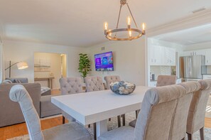Ein moderner Essbereich mit einem weißen Tisch, gepolsterten Stühlen, einer Chandelierleuchtre und einem Fernseher an der Wand.